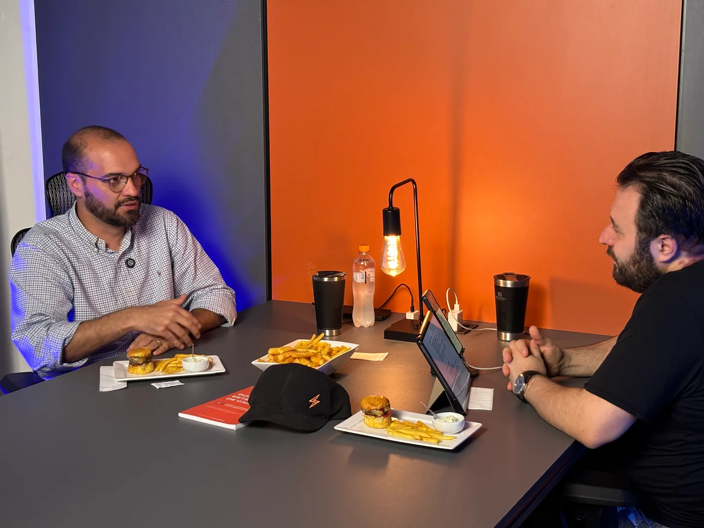Duas pessoas conversam à mesa do podcast, com microfones, iluminação e câmeras.